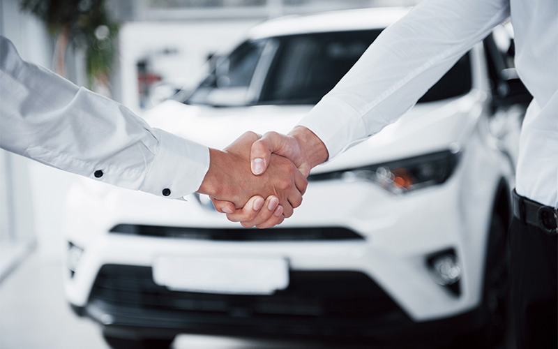 Two people shake hands in front of white car.